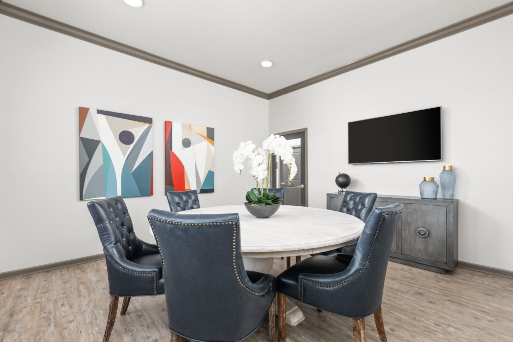 A five chair round table-set in a room of neutral tones with colorful framed artworks