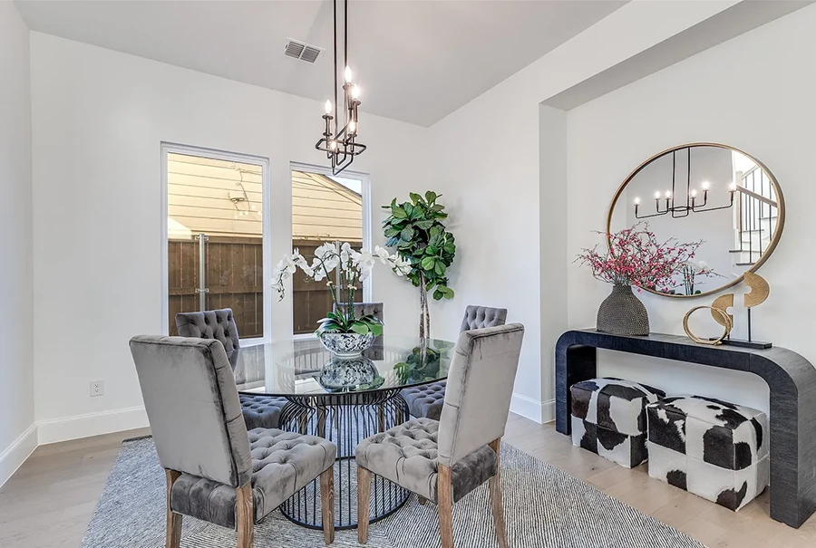 Dining area staged with a large round glass table, five grey velvet high back chairs, a black small console table, round gold mirror and two rawhide ottomans
