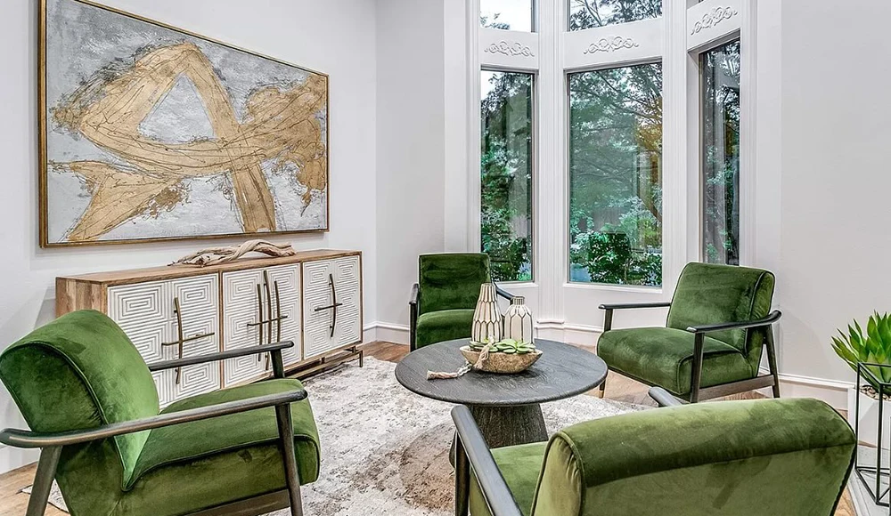 Seating area with four green velvet chairs, small round wooden coffee table, white and light wooden credenza, and large gold abstract painting