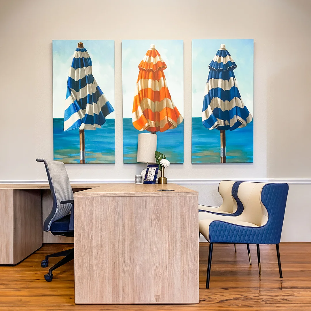 Office with light wood l-shaped desk, blue and white guest chairs and three beach umbrella paintings