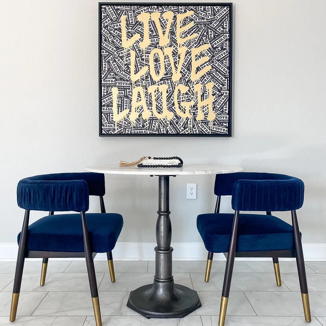 Small round white table, two dark blue velvet chairs and artwork that says live, love, laugh