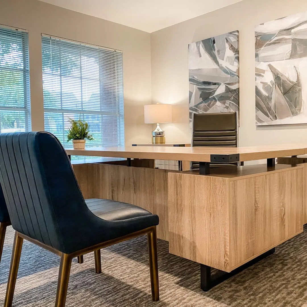 Leasing office with light wood colored l-shaped desk, two dark green leather charis and light colored abstract artwork